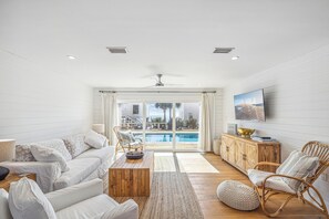 Una sala de estar moderna con un sofá blanco, una mesa de centro de madera, sillas de mimbre y vistas a una piscina a través de amplios ventanales.