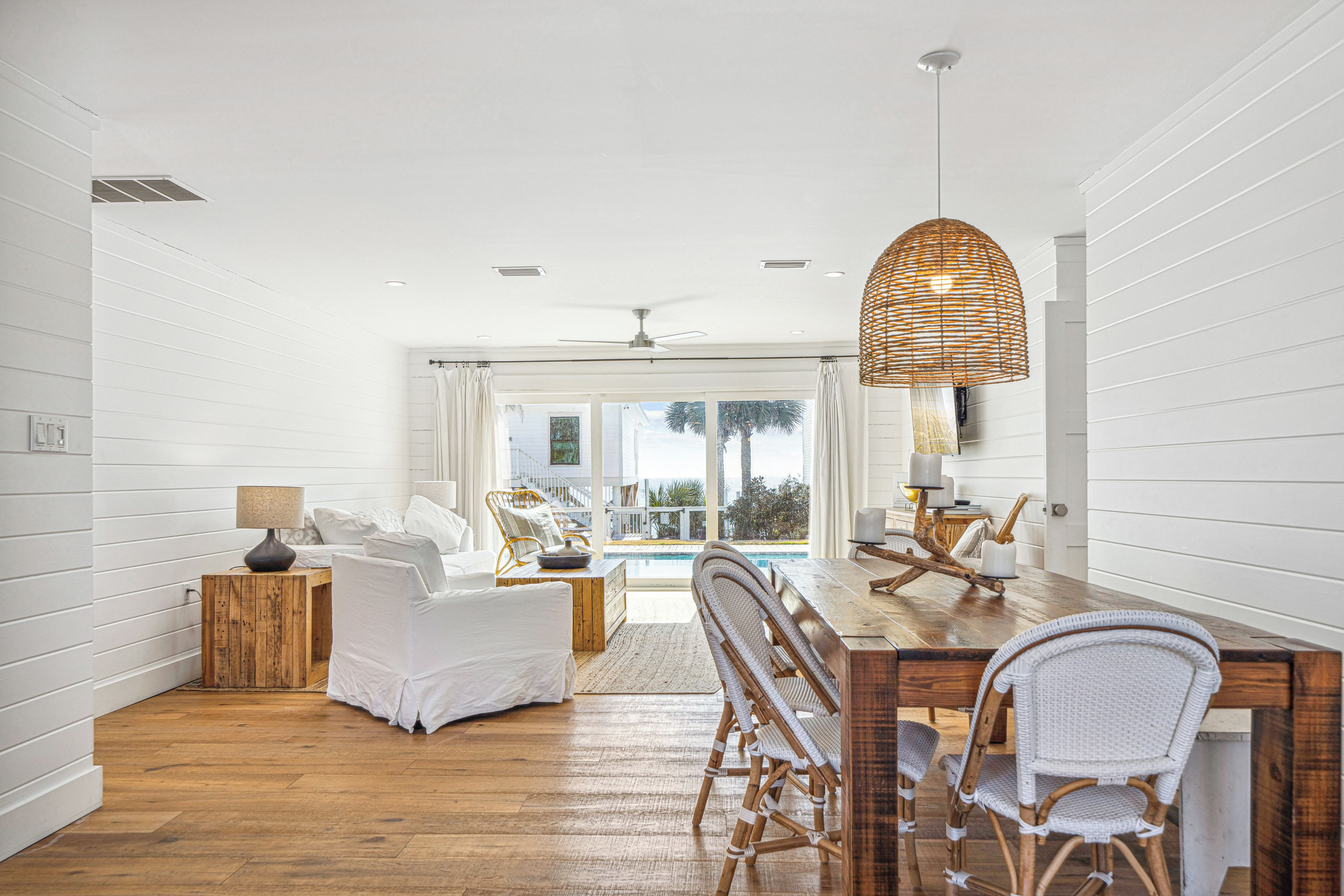 A spacious room with a wooden dining table, wicker chairs, and a large window overlooking a pool.