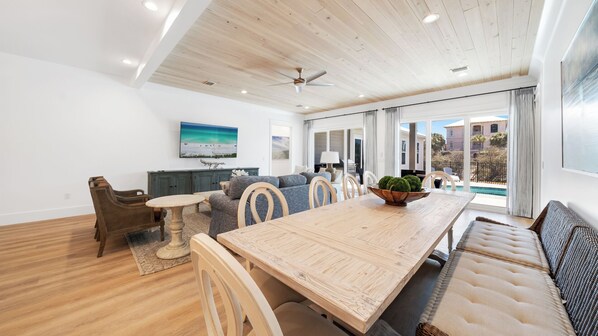 A spacious living area with a wooden dining table, a sofa, and a large window offering a view of the outdoors.