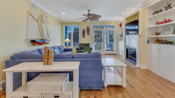 A living room with a sectional sofa, a white coffee table, wicker baskets, a ceiling fan, and a bookshelf with decorative items.