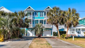 A multi-story beachfront house with a balcony, palm trees, and a clear blue sky.