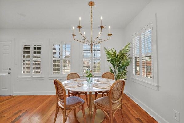 A dining room with a round table, six wicker chairs, a chandelier, and large windows with shutters.