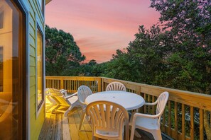 A wooden deck with a round table and four chairs, surrounded by lush greenery and a sunset sky. - OS758- 4 Bedrooms, Saltwater Pool & Hot Tub and 2 Spacious living areas (Corolla)