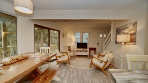 Un salon chaleureux avec une table en bois, des chaises blanches, un téléviseur et un escalier menant à un autre niveau.