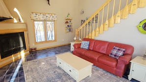 Uma sala de estar com lareira, um sofá vermelho, uma mesa de centro, uma escada com degraus de madeira e uma janela com cortinas estampadas.