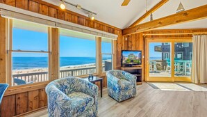 A room with large windows overlooking the beach, wooden flooring, two patterned armchairs, a television, and a ceiling fan. - Oceanfront Home in Avon with Hot Tub, Grill, Smart TVs. Pure Beachfront Bliss! (Avon)