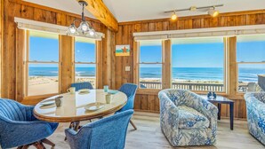 A room with a wooden interior, large windows with a beach view, a round table with chairs, and a patterned sofa. - Oceanfront Home in Avon with Hot Tub, Grill, Smart TVs. Pure Beachfront Bliss! (Avon)