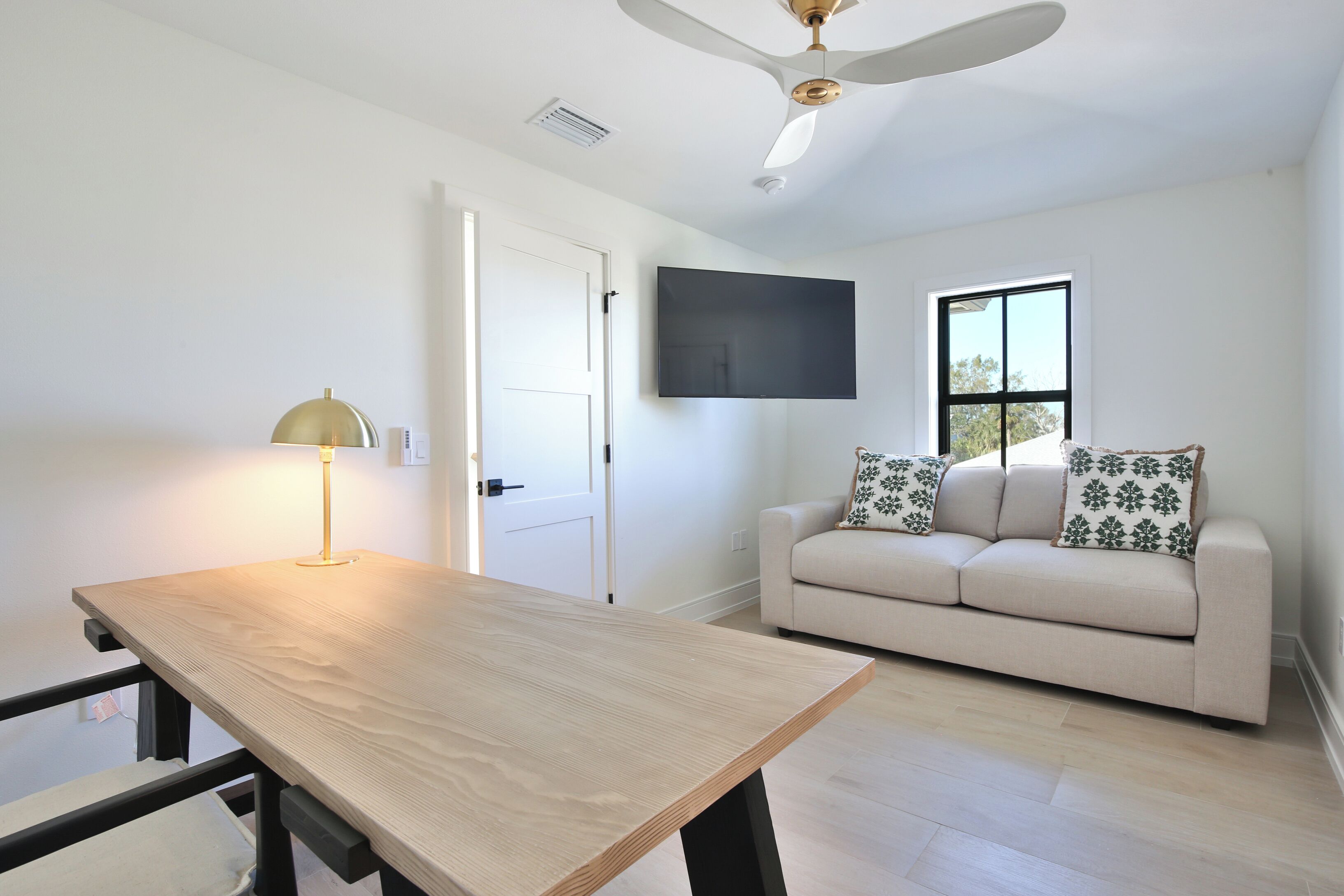 A room with a wooden table, a sofa with patterned cushions, a ceiling fan, and a mounted TV.
