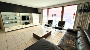 Ein modernes Wohnzimmer mit einer schwarzen Ledersofa, einem Glastisch und einem Flachbildfernseher auf einer weißen Kommode. Zwei schwarze Sessel und ein großes Fenster mit Vorhängen sind ebenfalls vorhanden.