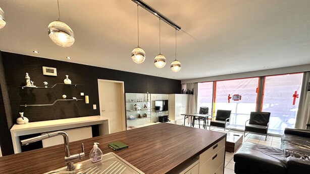 Eine moderne Küche mit Holzarbeitsplatte, Spüle aus Edelstahl und Pendelleuchten. Es gibt einen Essbereich mit Tisch und Stühlen sowie ein Wohnzimmer mit Sofa und Fernseher.