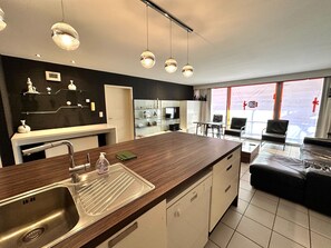 Une cuisine moderne dotée d’un plan de travail en bois, d’un évier en acier inoxydable et de luminaires suspendus. On y trouve un coin repas avec une table et des chaises, ainsi qu’un salon avec un canapé et un téléviseur.