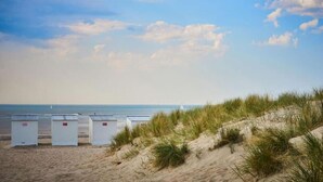 Ein Strand mit Dünen und drei weißen Bauten im Vordergrund, möglicherweise Strandkörbe.