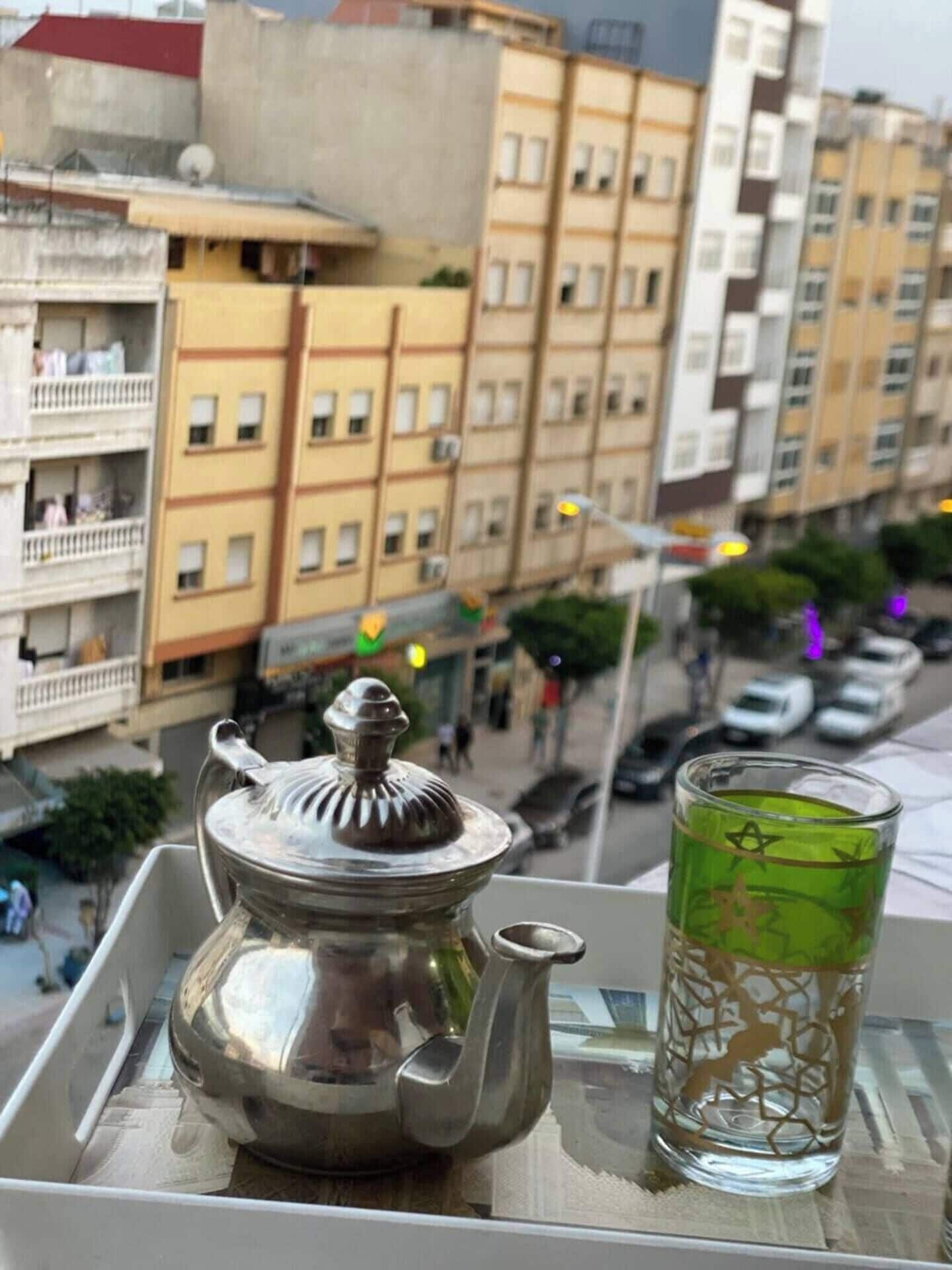 A tray with a metal teapot and a glass of tea, viewed from a window overlooking a cityscape with multi-story buildings.