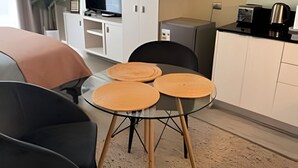 Ett modernt vardagsrum med ett glasbord, två stolar och en soffa. I bakgrunden finns ett köksområde med skåp och vitvaror.
