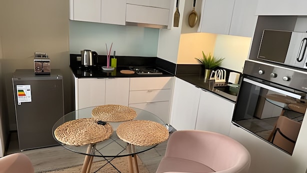 Ett modernt kök med vita skåp, en svart bänkskiva, ett glasbord med vävda bordstabletter och rosa stolar.