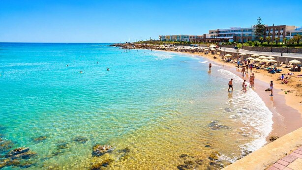 A beach with clear turquoise water, a sandy shore, and a row of buildings in the background. - Villa Amare - Heraklion Private Pool & Garden Gem (Kato Gouves)