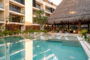 A pool with lounge chairs, a thatched-roof structure, and a multi-story building with balconies.