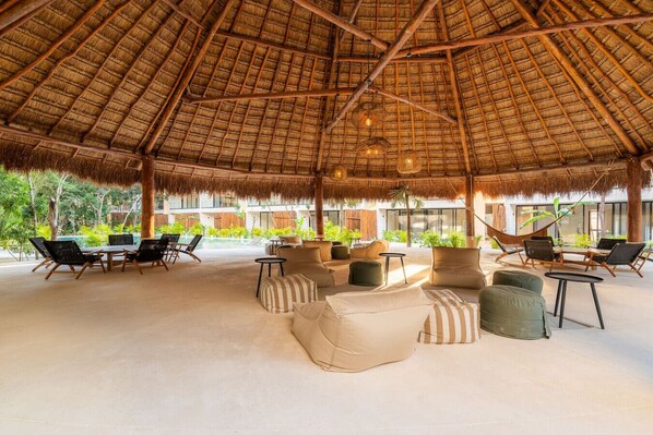 A thatched-roof outdoor lounge area with cushioned seating and tables, surrounded by greenery and a pool.