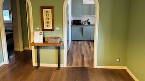 Un couloir au plancher de bois mène à une cuisine avec des meubles bleu clair et un four à micro-ondes. Un tableau encadré est accroché au mur et un panier est posé sur une petite table.