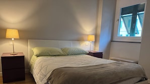 Une chambre à coucher avec un lit, deux tables de chevet avec des lampes, une fenêtre avec des stores et un plancher en bois.
