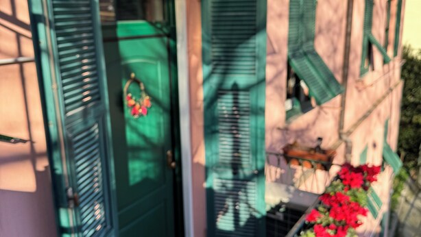 Un bâtiment à la façade rose, aux volets verts et doté d’un balcon orné de fleurs rouges.