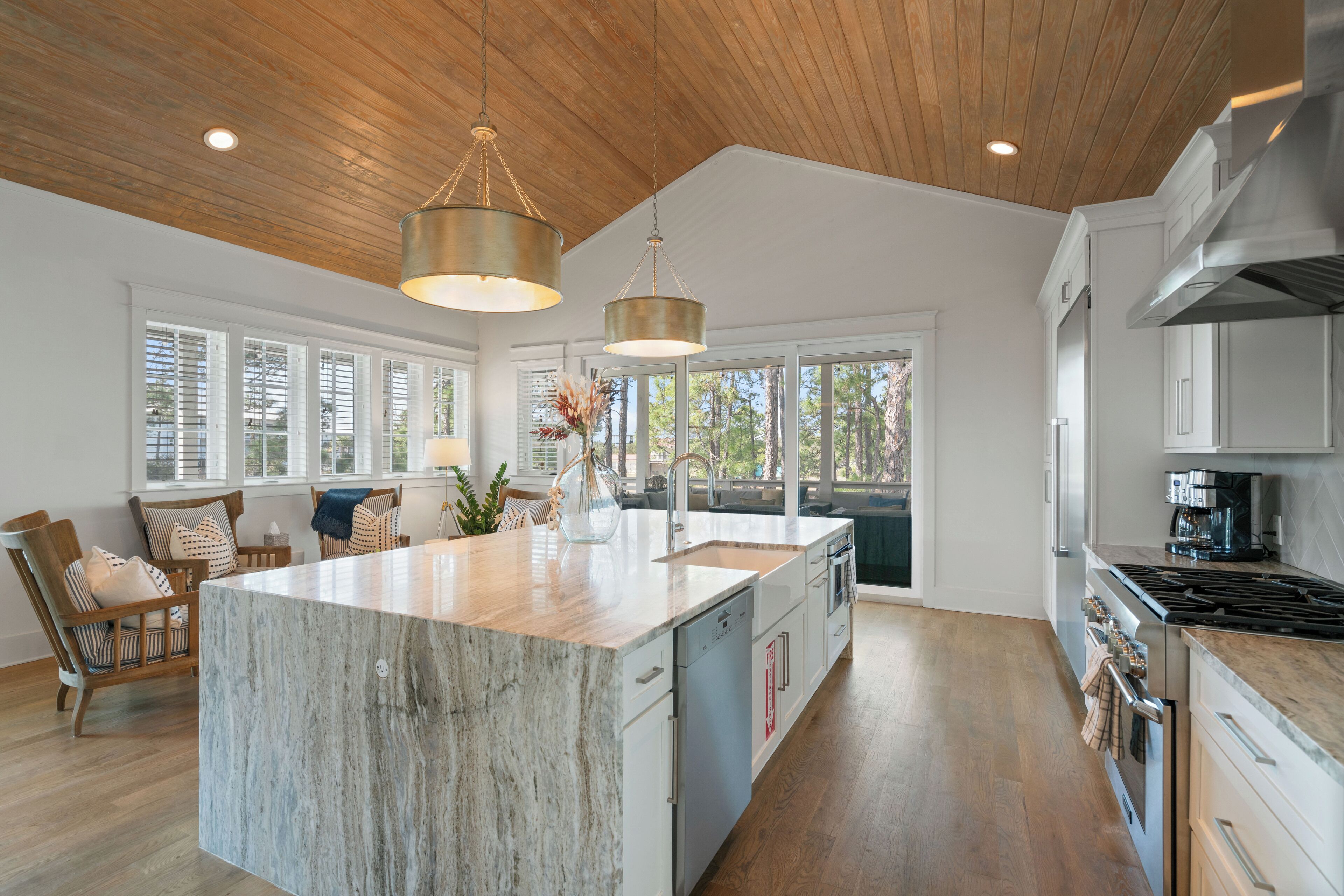 A spacious kitchen with a large island, wooden ceiling, and large windows offering views of the outdoors.