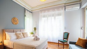 Un dormitorio con cama, una silla, un sofá, una ventana con cortinas y un equipo de aire acondicionado.