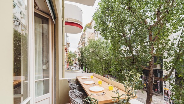 Un balcón con mesa de madera dispuesta para comer, rodeado de sillas, con vista a una calle con edificios y árboles.