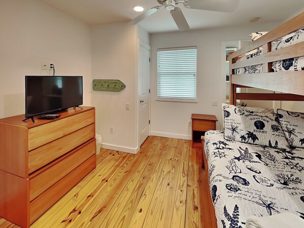 Een slaapkamer met een houten dressoir, een televisie, een stapelbed met beddengoed met een zeemeer thema, een raam met jaloezieën en een plafondventilator.