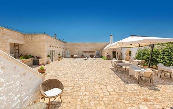 Un patio exterior con suelo de piedra, sillas de mimbre y mesas bajo sombrillas.