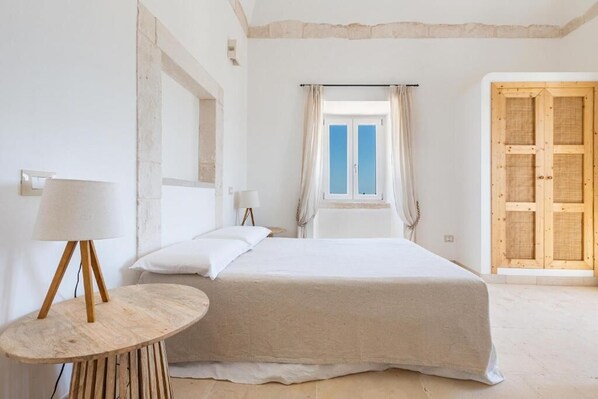 Un dormitorio con cama, una mesa redonda de madera con lámpara, una ventana con cortinas y una puerta de madera.
