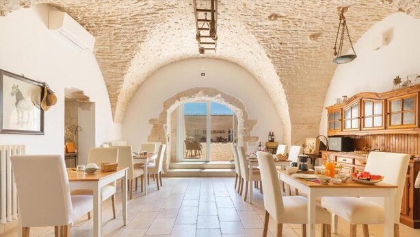 A dining area with white chairs and tables, stone walls, and a view through an arched doorway.
