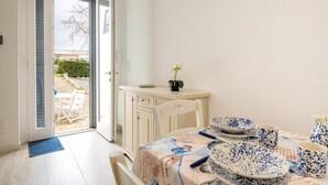 A room with a dining table set with blue and white patterned plates and cups, a white chair, a small cabinet with a potted plant, and a door leading to an outdoor area. - The Villa Resort, sleeps 2 + shared pool (MARTINA FRANCA)