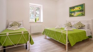 A room with two single beds, each with a green bedspread and white headboard. There is a window with a view of greenery outside, and a painting of a tropical landscape on the wall. - The Villa Resort, sleeps 8 + shared pool (MARTINA FRANCA)