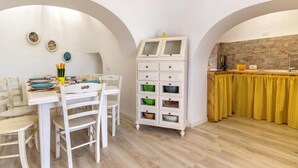 A dining area with a white table and chairs, a white cabinet with multiple drawers, and a kitchen area with a stone backsplash and yellow curtains. - The Villa Resort, sleeps 8 + shared pool (MARTINA FRANCA)