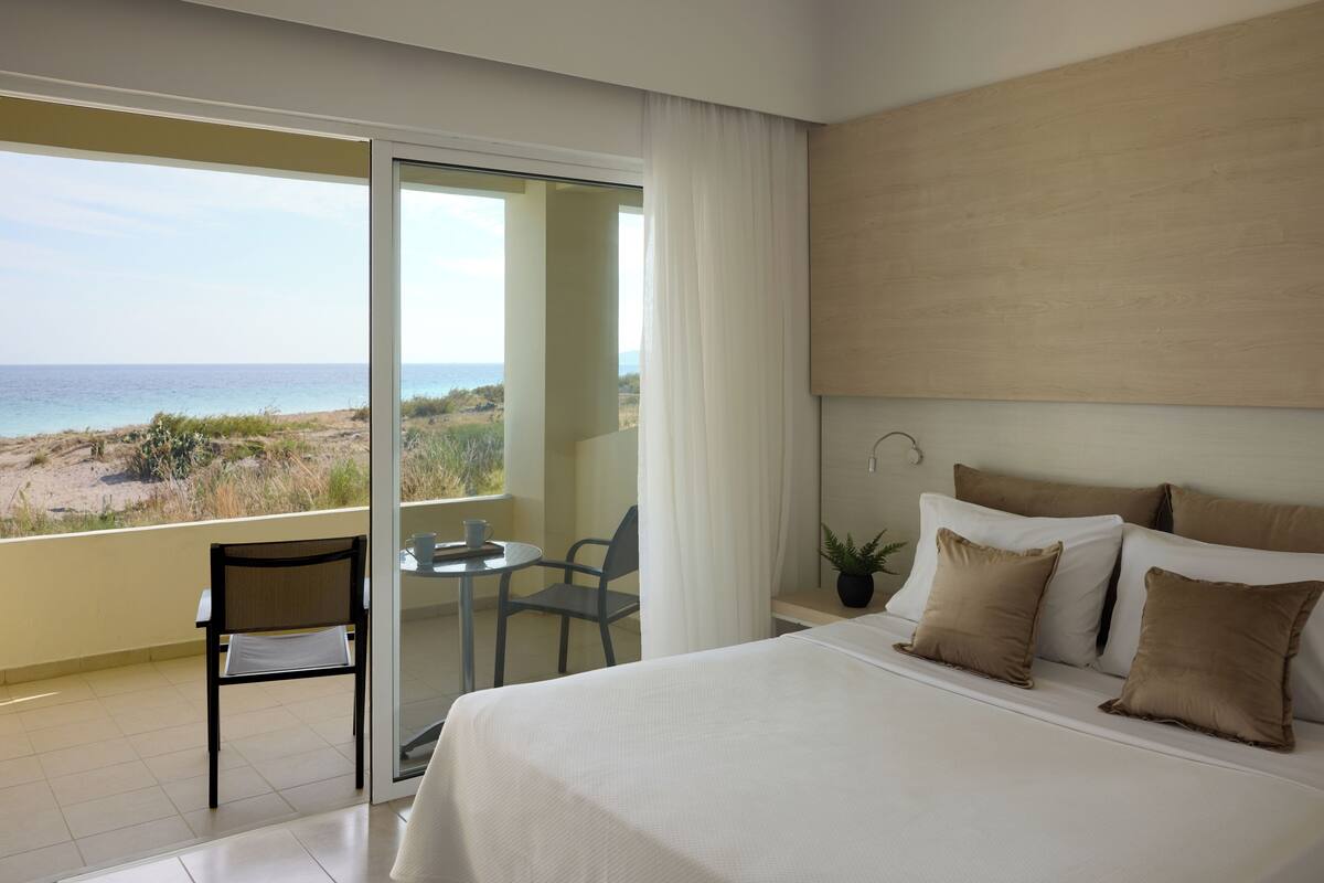 A hotel room with a bed, a chair, a small table, and a view of the beach.