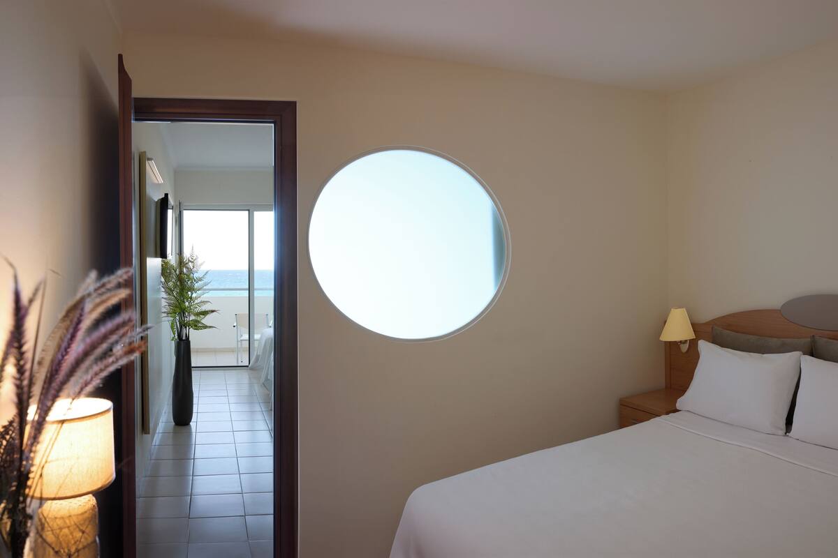 A hotel room with a large round mirror, a bed with white linens, a nightstand with a lamp, and a view of the beach through an open door.