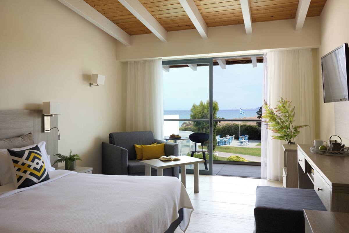 A modern hotel room with a large bed, a sofa, a small table, and a balcony with a view of the sea.