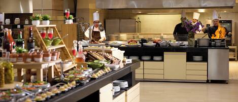 Een moderne keuken met koks die eten bereiden, een buffet met diverse gerechten, en een netjes, overzichtelijk interieur.