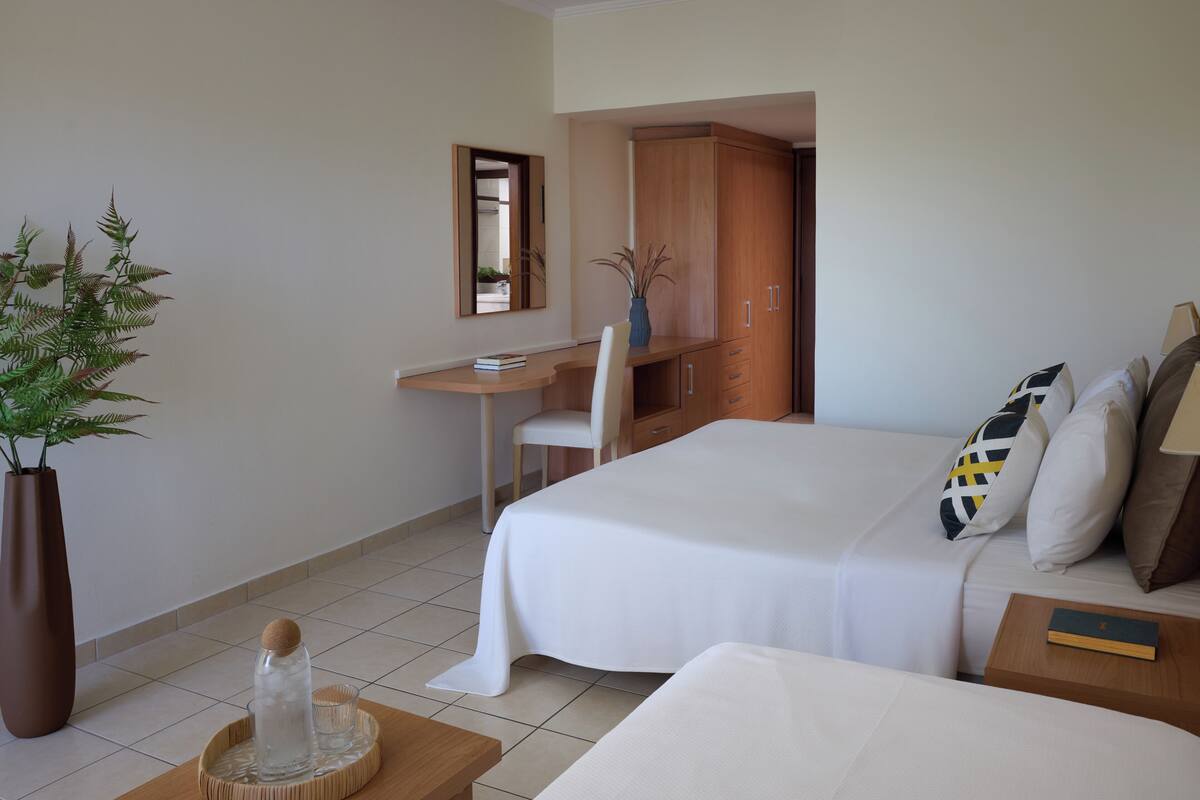 A hotel room with a bed, a desk, a chair, a large potted plant, and a small table with a tray.