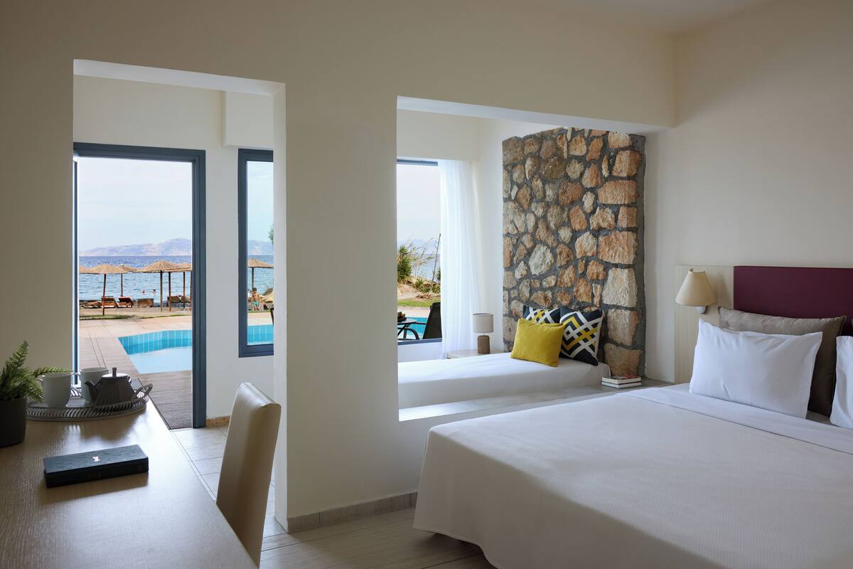 A hotel room with a bed, a desk with a plant, a stone wall, and a view of a pool and beach.