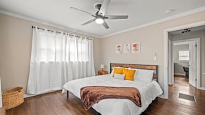 Quarto com cama, ventoinha no teto, mesa de cabeceira, abajur e porta que dá para outro cômodo.