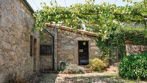 Un edificio in pietra con tetto in tegole, una panca di legno e un’area giardino con piante e una recinzione.