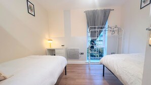 Un dormitorio con dos camas, una mesita de noche con lámpara, un radiador y una ventana con cortinas.