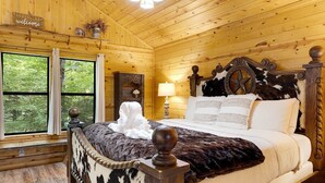 Un dormitorio de cabaña de madera con cama, mesita de noche, lámpara y ventana con vista a árboles.