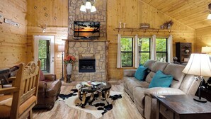 Interior acogedor de una cabaña con chimenea de piedra, un sofá de cuero, una mesa de comedor de madera y un amplio ventanal con vistas al exterior.