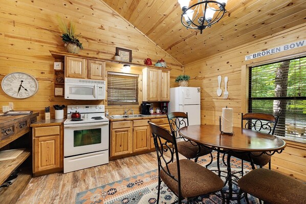 A cozy cabin-style kitchen with wooden cabinets, a stove, a microwave, and a refrigerator. There is a dining table with chairs and a rug on the floor.