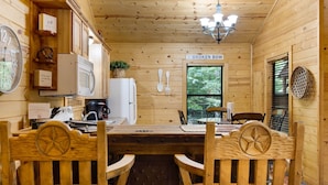 Interior de una cabaña de madera con una mesa de comedor, sillas, una zona de cocina, un letrero que dice “BROKEN BOW” y un ventanal grande.