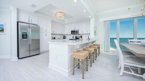 Una cocina moderna con armarios blancos, un refrigerador de acero inoxidable, una isla central con taburetes y vistas al océano a través de amplias ventanas.