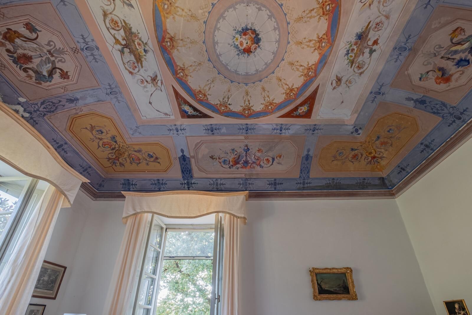Una habitación con un techo decorativo que presenta motivos florales y geométricos, un ventanal con cortinas y cuadros en las paredes.